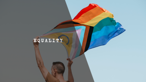 A person holding up a large LGBTQ+ Progress Pride flag waving against a clear sky, symbolizing the fight for visibility and equality.
