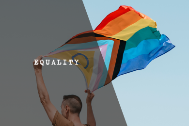 A person holding up a large LGBTQ+ Progress Pride flag waving against a clear sky, symbolizing the fight for visibility and equality.