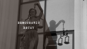 A black and white, high-contrast image depicting the statue of Lady Justice holding her scales, with her shadow cast on the wall behind her resembling the silhouette of a corrupt or grubby man to symbolise judicial capture.