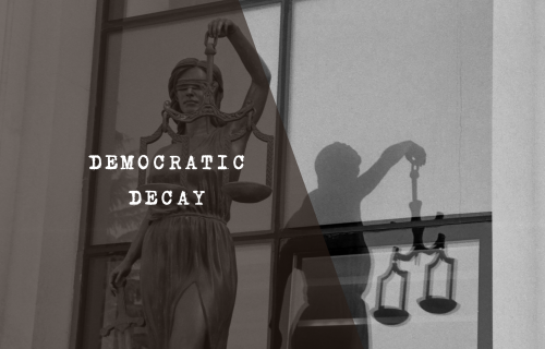 A black and white, high-contrast image depicting the statue of Lady Justice holding her scales, with her shadow cast on the wall behind her resembling the silhouette of a corrupt or grubby man to symbolise judicial capture.